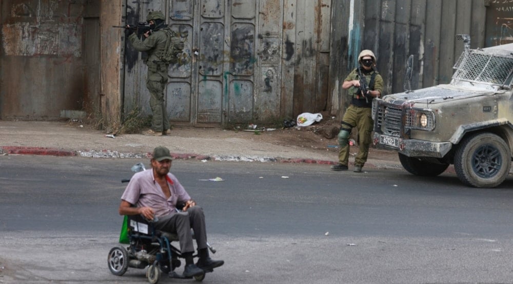 جنود الاحتلال الإسرائيلي يراقبون رجل فلسطيني يستخدم كرسياً متحركاً يمر من جانب آلياتهم في أثناء قيامهم بمداهمة في مدينة نابلس في الضفة الغربية (أ ف ب)